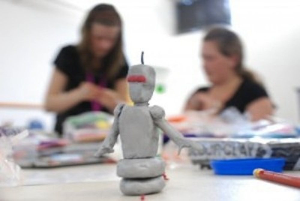 Photo of two young people in the background and a plasticine model of a robot in the foreground