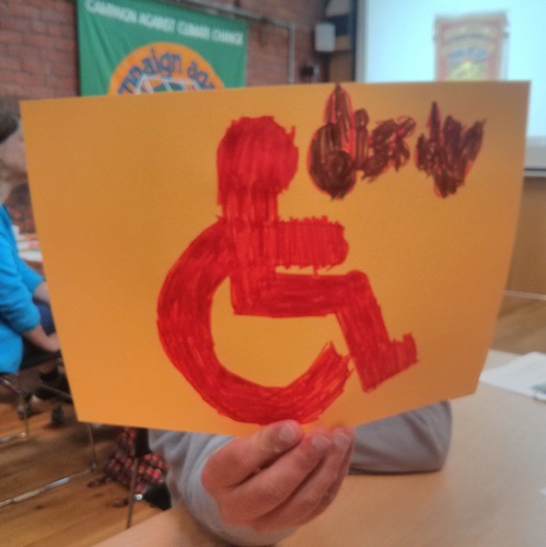 participant to the protest banner workshop holding up to the camera his banner idea of the wheelchair accessible sign