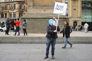 image of a man holding a sign saying 'alone again. he's wearing a mask with the facebook like symbol on