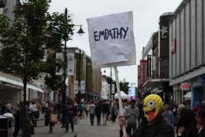 man holding up a sign saying 'empathy'. he has a mask on with the smiley face Emoticon on it