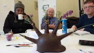 a photo of three people sat around a table. In the centre of the table is a hat with spikes on it, each spike holds a flower