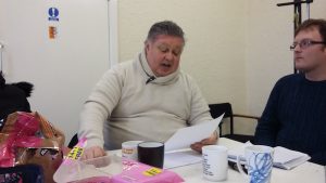 photo of people sat around the table one man is reading from a piece of paper