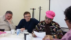 people sat a round a table a lady in a pink hat reads from a piece of paper