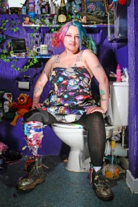 image of a woman sitting on a toilet. she has colourful hair and a prosthetic leg